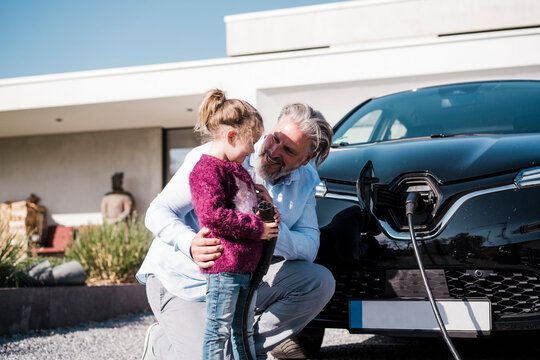 Happy man with daughter holding electric plug in front of car