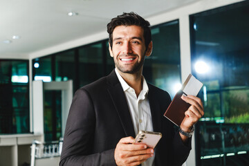 Smiling businessman holding smartphone, passport, and boarding pass at the airport. Concept of air travel, check-in service, and professional global mobility with modern digital convenience.