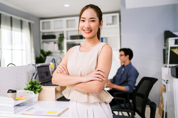 Confident Asian businesswoman smiling with arms crossed in modern bright office. Male colleague working in background. Teamwork, diversity, and success in a professional environment.