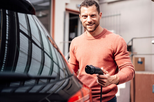 Smiling man holding charging cable standing by electric car