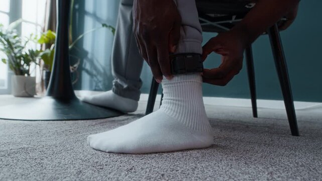 Cropped shot of hands of unrecognizable African American man under house arrest sitting at table and adjusting electronic monitoring bracelet on his ankle