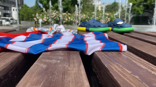 A vibrant red, white, and blue striped shirt resting on a rustic wooden bench, with blue and yellow/green children's clogs in the background, set against a backdrop of splashing fountain jets