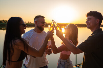 Four cheerful friends are toasting with beer bottles on a lake pier at sunset, enjoying the golden...