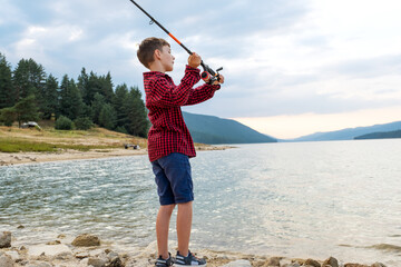 Red plaid shirt boy fishing calm clear lake surrounded forest hills enjoying rural summer vacation...