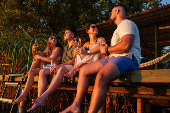 Four friends are relaxing on a wooden pier, enjoying refreshing drinks as they watch a beautiful sunset over a tranquil lake, creating a perfect summer evening