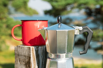 Travel morning with moka coffee pot and red mug, peaceful lake view, outdoor caffeine ritual in wilderness destination.Silver Italian-style espresso pot next to retro red mug, nature-inspired backdrop