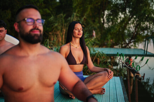 Group of friends meditating sitting in lotus position on a pier, enjoying the sunset over the lake, finding inner peace and well being through yoga practice