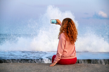 Seaside adventure as young woman records wave explosion on smartphone, pastel horizon and marine force collide.. Enjoy beauty of sea sunset. Relax. Lifestyle