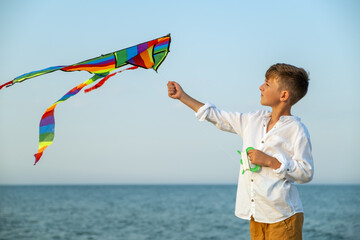 Close-up of boy in summer clothes flying rainbow kite near calm sea, simple outdoor joy, blue sky...