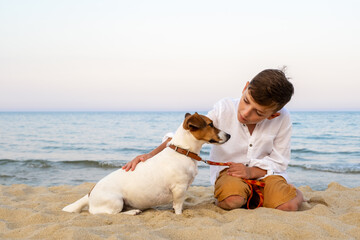 Boy sitting on sand with Jack Russell terrier, sunset beach moment full of affection and calm,...