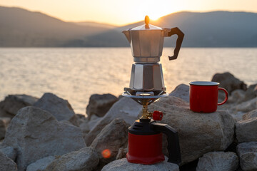 Red metal cup and moka pot on portable stove against peaceful lake and hills background at dusk,...