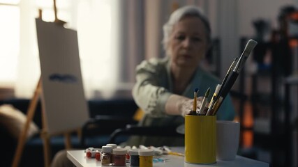 Senior lady artist in wheelchair painting on canvas using easel, art therapy