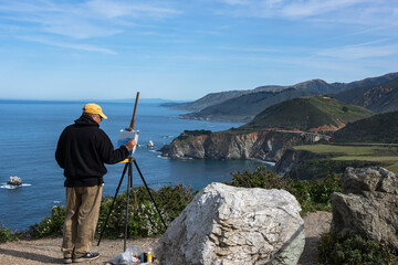 An artist stands at an easel painting the landscape of the California coast.