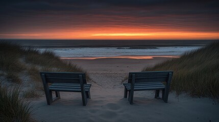 Two dark benches face the ocean at sunrise.  A dramatic sunrise colors the sky.  Sandy dunes flank the benches.  Ocean waves gently lap the shore