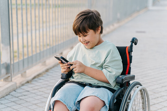 Smiling teenager boy in a wheelchair using cell phone for call, vieo chat or gaming, technology lifestyle concept