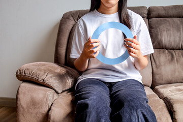 Woman Sitting On Couch Holding A Large Blue Circle Symbol At Home, Representing Diabetes Awareness