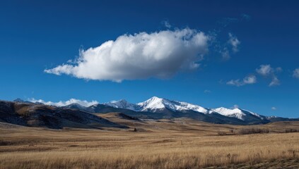 Snow-capped mountains rise above a vast, dry, autumnal plain under a clear blue sky, a single cumulus cloud floats serenely