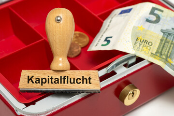 Ein Holzstempel ist beschriftet mit dem Aufdruck Kapitalflucht und liegt symbolisch in einer leeren...