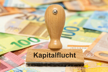 Ein Holzstempel ist beschriftet mit dem Aufdruck Kapitalflucht und steht über vielen Euro Banknoten