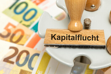 Ein Holzstempel ist beschriftet mit dem Aufdruck Kapitalflucht und steht über vielen Euro Banknoten