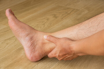 Person pressing sore ankle above heel, showing foot pain or strain. Barefoot on wooden floor in...