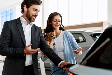 Car salesman showing electric vehicle charging to woman holding chihuahua