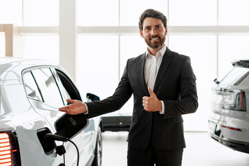 Car salesman showing charging electric car and giving thumbs up in dealership