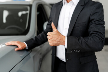 Car salesman showing thumbs up while touching new car in dealership
