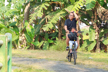 asian father and elementary-age son enjoying riding a bike outdoors in a park.