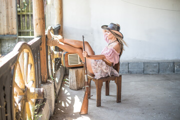 Beautiful young woman dressed in western cowgirl style, wearing a hat and boots, sitting on a chair...