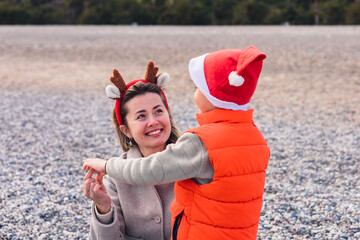 A smiling Asian woman with reindeer antlers interacts with her young son wearing a Santa hat and orange vest on a winter beach during the New Year.