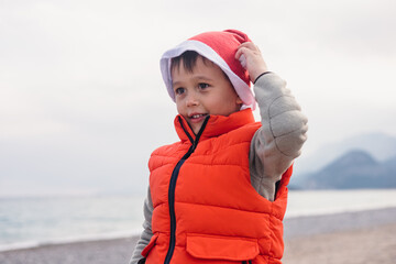 A young boy wearing an orange vest and a Santa hat stands on a beach during winter. The ocean and mountains are visible in the background.