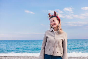 A young Caucasian woman stands on a beach during winter. She wears a beige jacket and festive reindeer antlers, smiling against a backdrop of the ocean and sky.