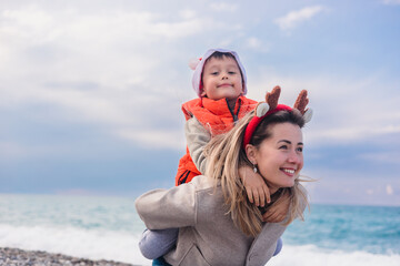 A young Caucasian woman with long blonde hair carries a smiling Asian boy on her back at the beach during winter. They wear festive clothing for New Year and Christmas.