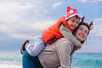 A young Caucasian woman with brown hair carries her smiling son on her back at the beach during winter. The boy wears a Santa hat and an orange vest.