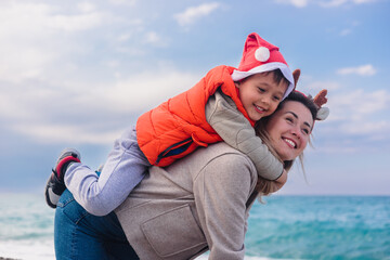 A young Caucasian woman carries her smiling son on her back at the beach during winter. The boy wears a Santa hat and a bright orange vest. The ocean is visible in the background.