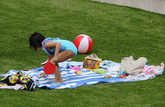 Ni&ntilde;a con ba&ntilde;ador azul, raqueta roja de ping pong y bal&oacute;n de playa jugando sobre la toalla a rayas en la hierba de la piscina de verano