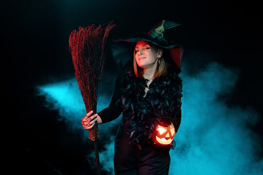 Young woman wearing halloween witch costume and hat on black background with magical blue smoke holds broom and pumpkin jack o lantern. Halloween party, copy space. - Powered by Adobe