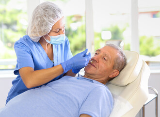 Obraz premium Adult female doctor in medical uniform makes injection in lips of elderly man in medical office