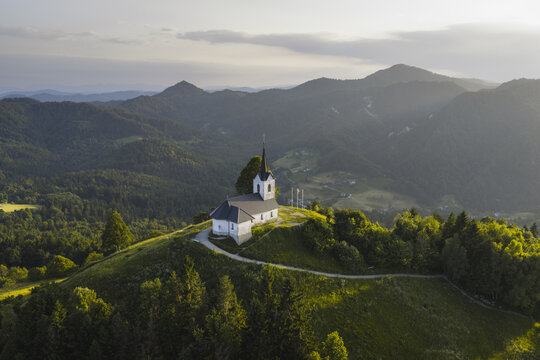 Aerial view of a church steeple piercing the skyline, bathed in the warm glow of the sun, amidst rolling hills and lush greenery, Sv Jakob, Ljubljana, Slovenia.