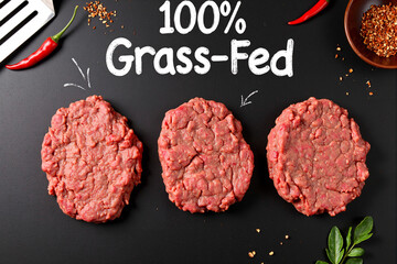 Three grass-fed ground beef patties on dark slate with 100% label and chili peppers, premium organic meat products for healthy burger cooking preparation