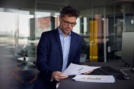 Businessman wearing eyeglasses reading document standing at desk in office