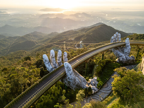 Aerial view of the iconic Golden Bridge, held by giant stone hands, stretching across the lush, sun-kissed mountains, DaNang, Da Nang, Vietnam.