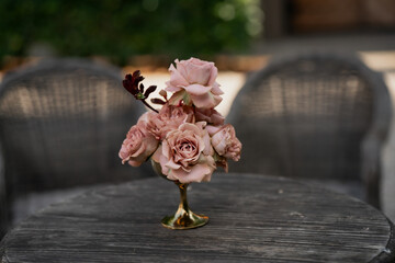 Vintage rose flower arrangement in elegant golden vase on rustic wooden table with soft chairs in...