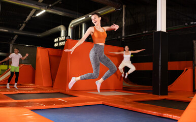 Enthusiastic middle-aged woman experiencing thrill of leaping high at vibrant trampoline gym with...