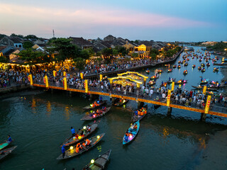Aerial view of boats adorned with lanterns glide along the river beneath bridges illuminated with golden lights, casting a warm glow on the ancient town, Há»™i An, Quáº£ng Nam, Vietnam.