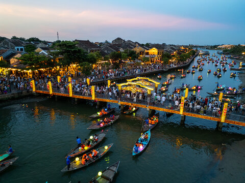Aerial view of boats adorned with lanterns glide along the river beneath bridges illuminated with golden lights, casting a warm glow on the ancient town, Há»™i An, Quáº£ng Nam, Vietnam.