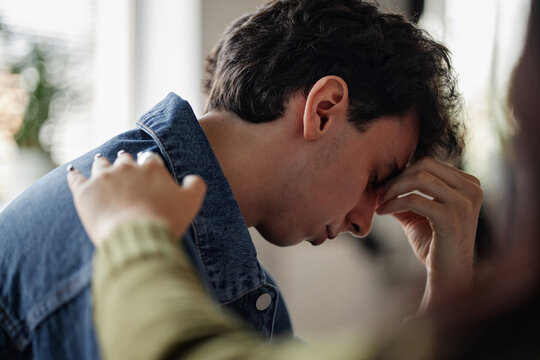 Caucasian teenage boy sitting with head in hand showing distress during counseling session, another placing supportive hand on his shoulder offering comfort and reassurance
