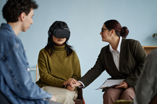Multiethnic group of teenagers attending counseling session, Asian teenage girl wearing virtual reality headset holding hands with female counselor, Caucasian teenage boy observing interaction