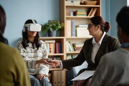 Diverse group of teenagers attending counseling session, teenage girl wearing virtual reality headset interacting with female counselor, other teenagers sitting in background